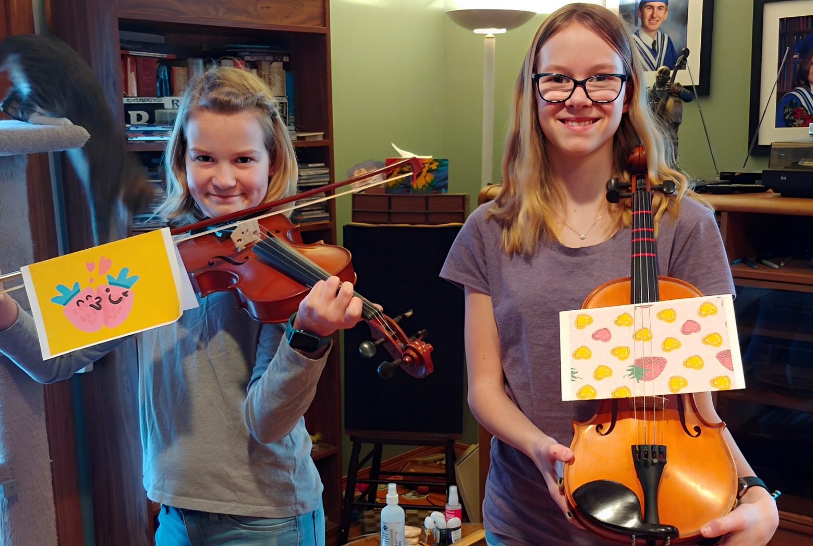 Two young girls holding violas indoors, each displaying colorful, handmade cards attached to their instruments. Shelves and a lamp are visible in the background.